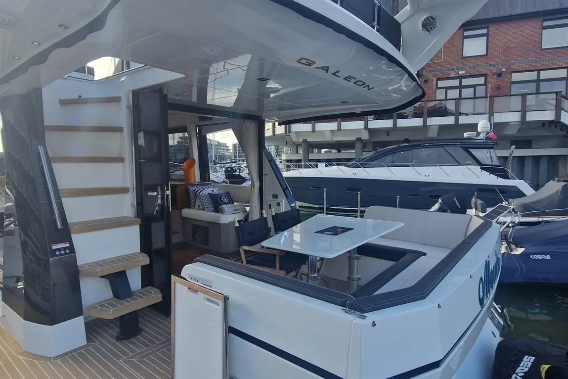 A modern yacht interior featuring steps to upper deck, lounging area with cushions, and a dining table, moored at a marina.